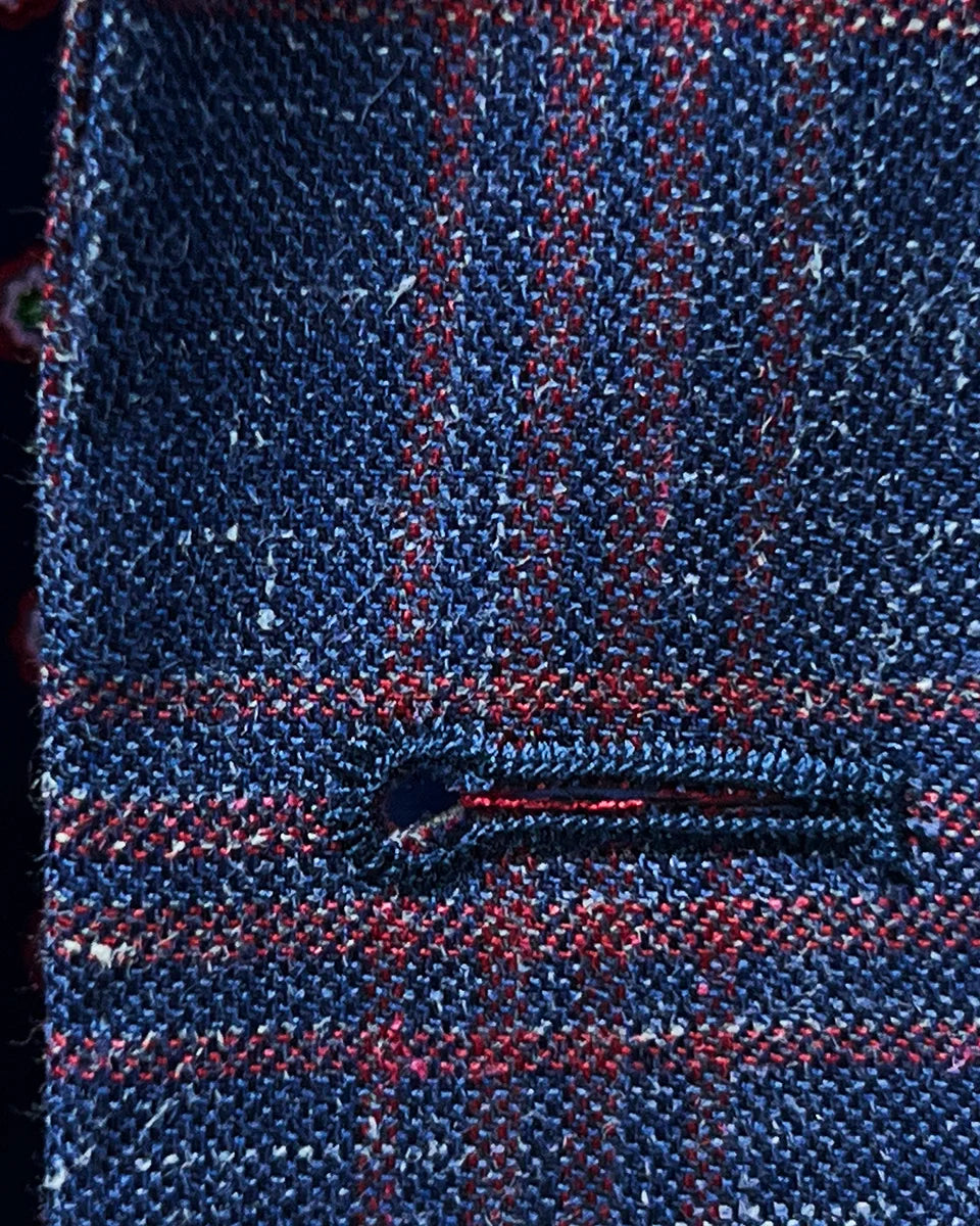 Macro view of hand-finished buttonhole stitching in dark berry maroon thread against indigo blue windowpane fabric background