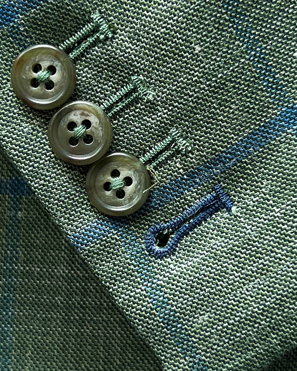Working sleeve buttonholes with royal blue contrast stitching on olive green windowpane fabric, showing four-button surgeon's cuffs