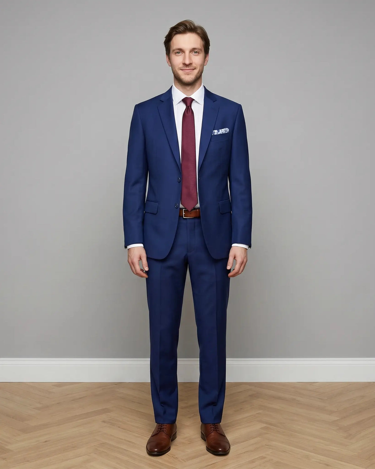 Full body front studio portrait of royal blue sharkskin men's suit showing tailored jacket, trousers, belt, burgundy silk tie, and brown leather shoes.