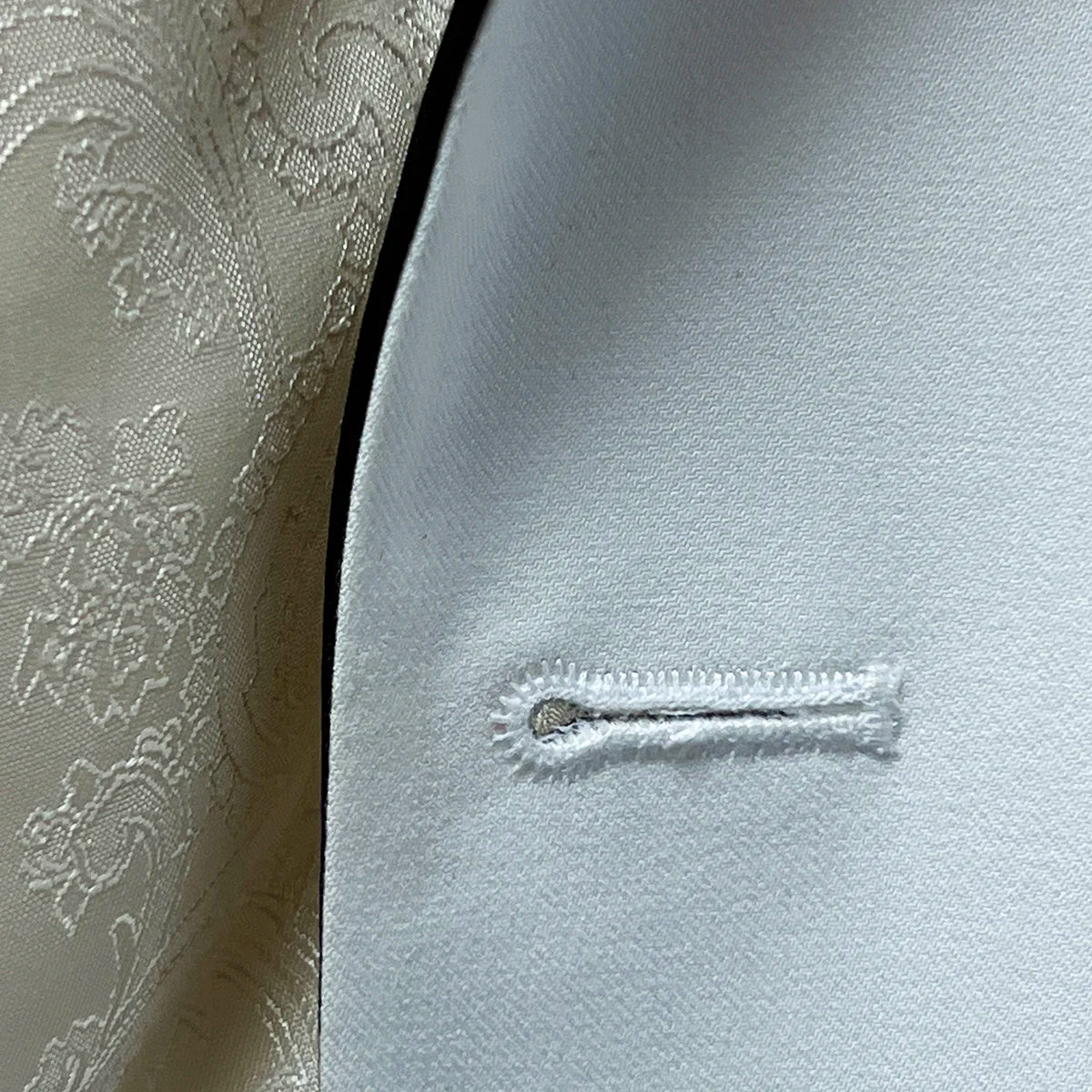 Detailed view of the buttonhole stitching on the ivory tuxedo jacket, emphasizing the fine tailoring and black satin accents.