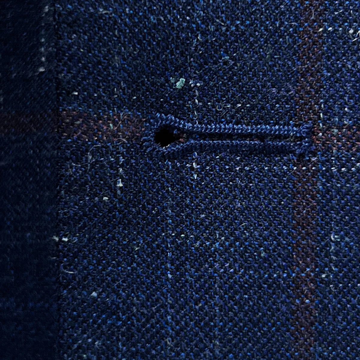 Macro view of hand-picked buttonhole stitching detail showing ruby red contrast thread work on navy windowpane fabric
