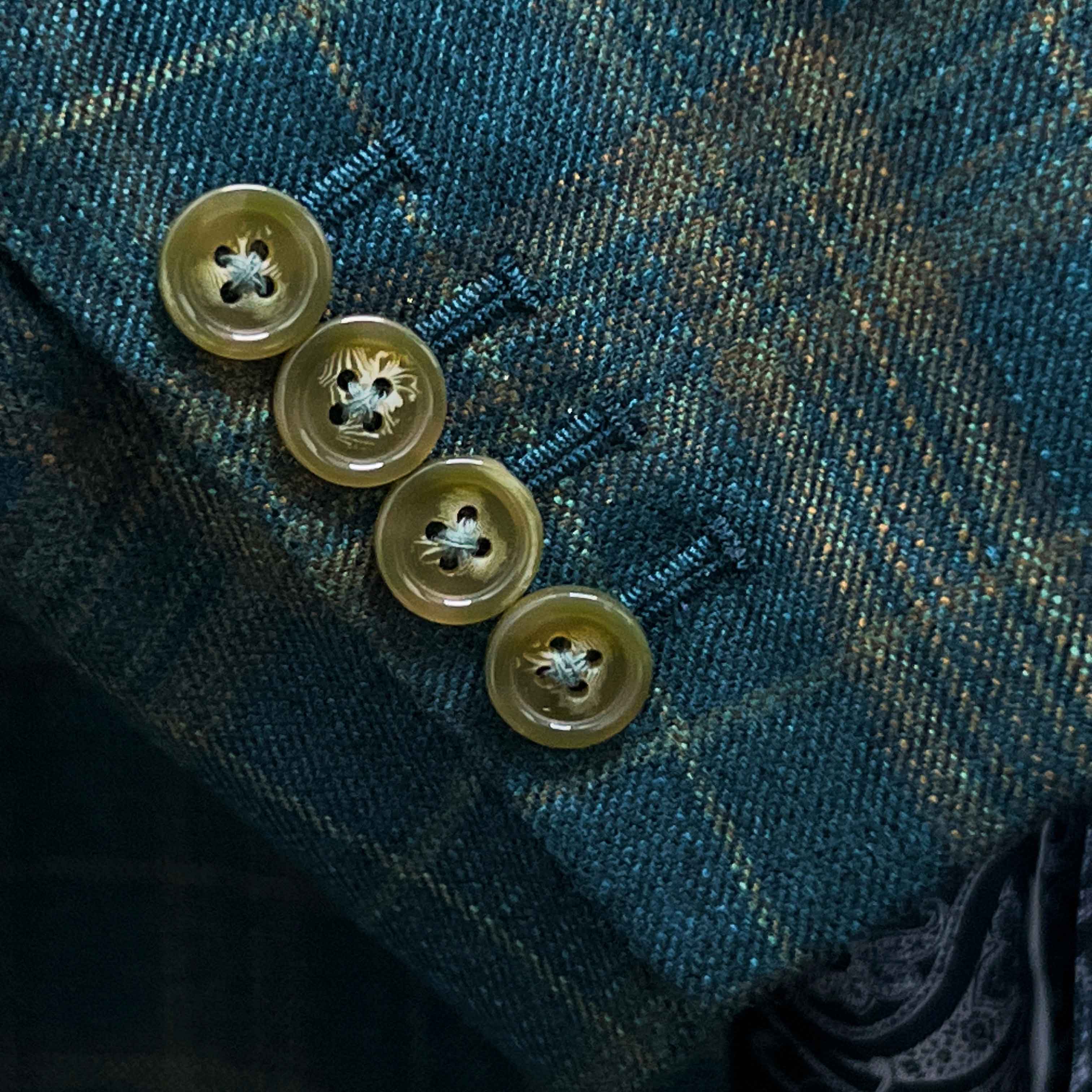 Suit jacket sleeve cuff showing functional buttonholes with contrast olive green accent stitching on deep teal green multi check fabric