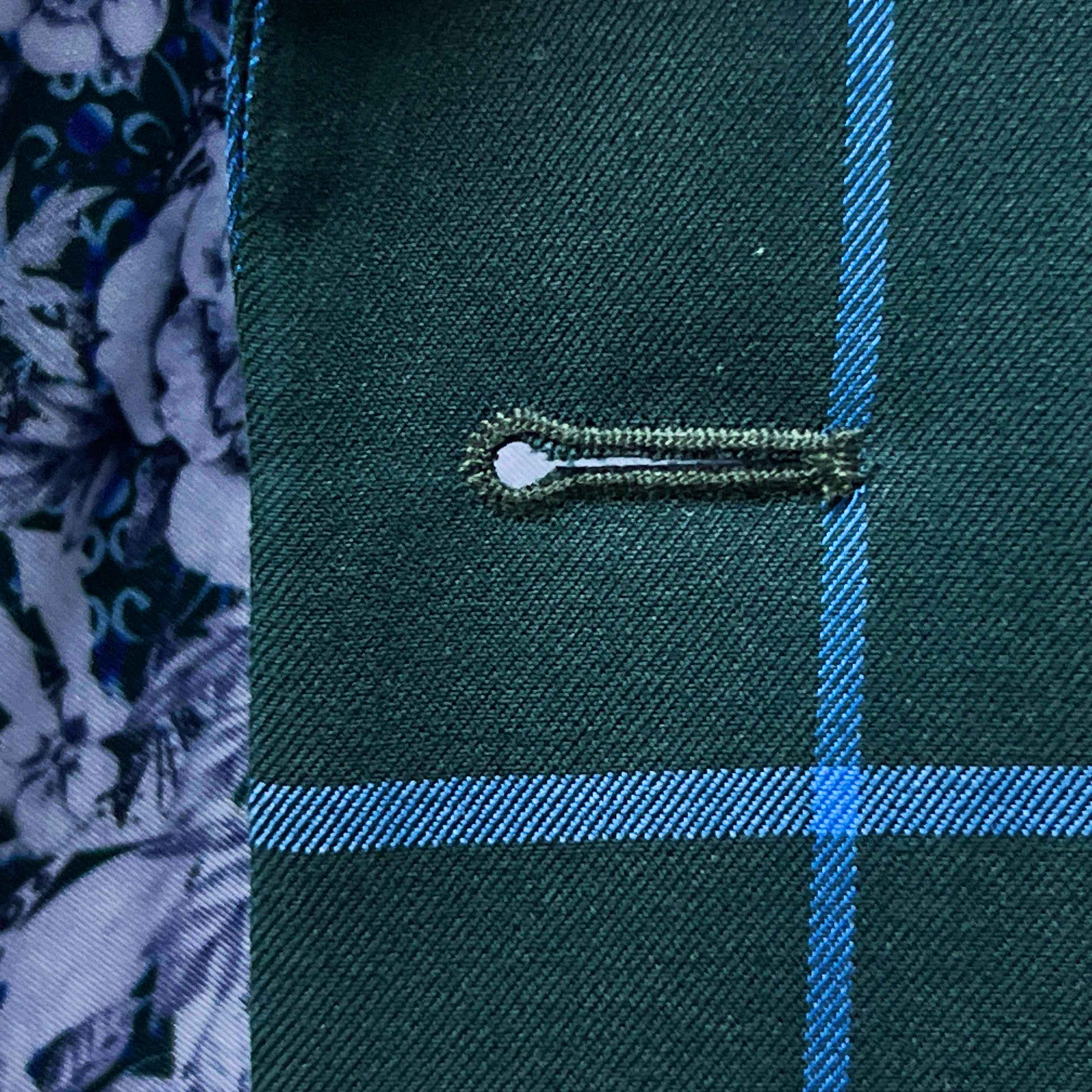 Detailed macro shot of hand-finished buttonhole stitching on forest green windowpane sport coat with bright blue contrast thread