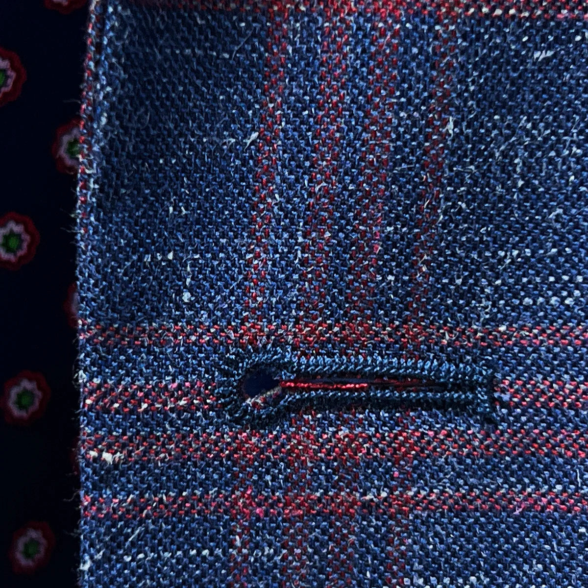 Macro view of hand-finished buttonhole stitching in dark berry maroon thread against indigo blue windowpane fabric background