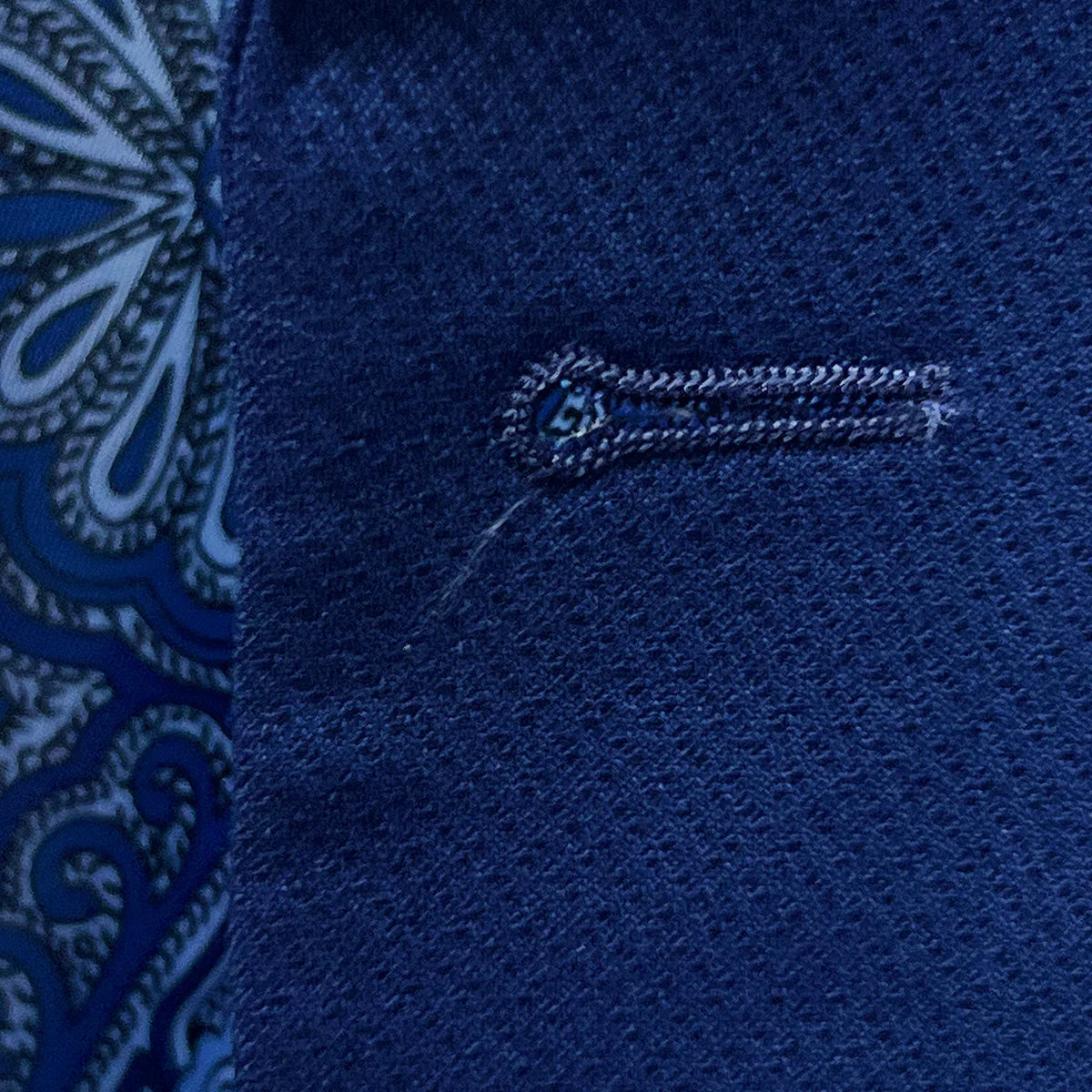 Detailed macro shot of hand-finished buttonhole stitching on navy blue geometric textured suit with light blue contrast thread