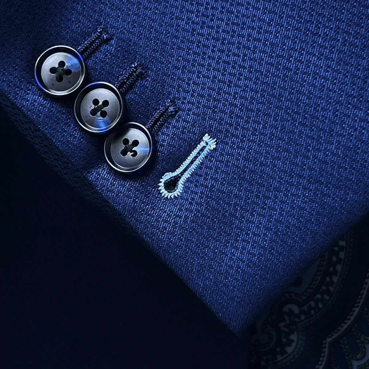 Suit jacket sleeve cuff showing functional buttonholes with contrast light blue accent stitching on navy blue geometric textured fabric
