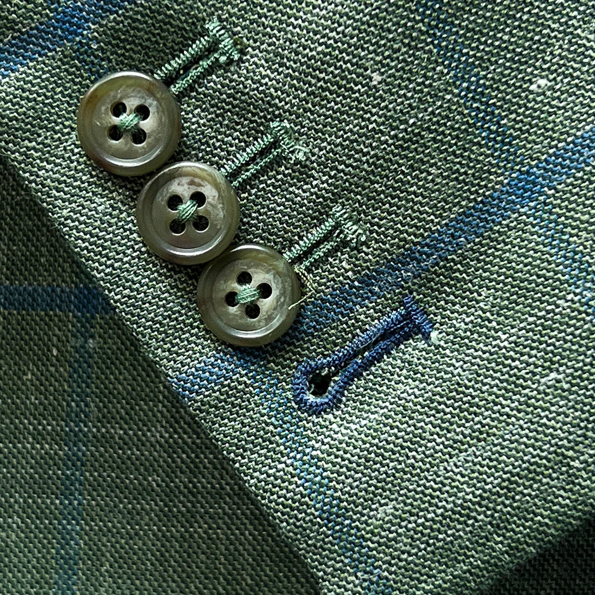 Working sleeve buttonholes with royal blue contrast stitching on olive green windowpane fabric, showing four-button surgeon's cuffs
