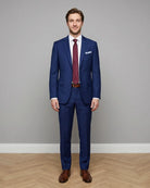 Full body front studio portrait of royal blue sharkskin men's suit showing tailored jacket, trousers, belt, burgundy silk tie, and brown leather shoes.